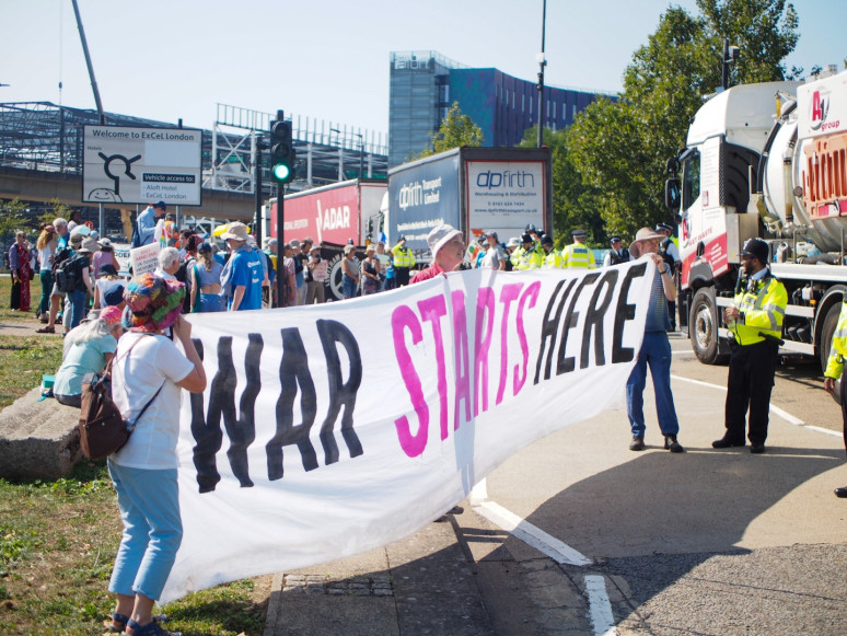 Demonstrators outside DSEI hold banner reading war starts here as large lorries deliver death products to the ExCel centre