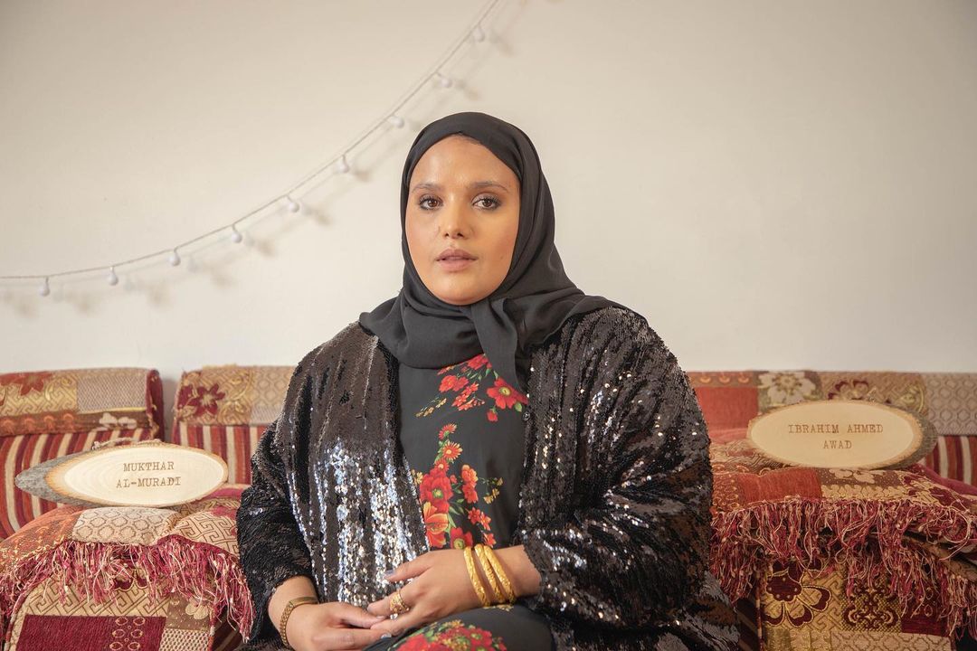 Poet Amina Atiq is sitting on a sofa, wearing a black outfit with a black headscarf. There are wooden plaques on both sides of her, reading the names of two casualties of the war in Yemen. The one on her right reads Ibrahim Ahmed Awad, and the one on the left reads Mukthar Al-Muradi