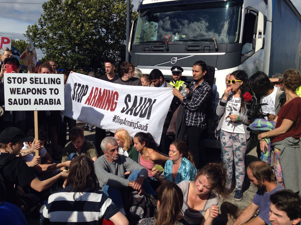 Join the Festival of Resistance at DSEI (crowd of campaigners block military equipment on the road, with Stop Arming Saudi banner