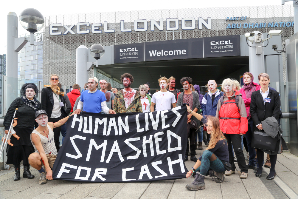 About 20 protesters stand in front of entrance to ExCeL London, with banner saying Lives smashed for cash. Some are made-up with bloody faces and injuries. 