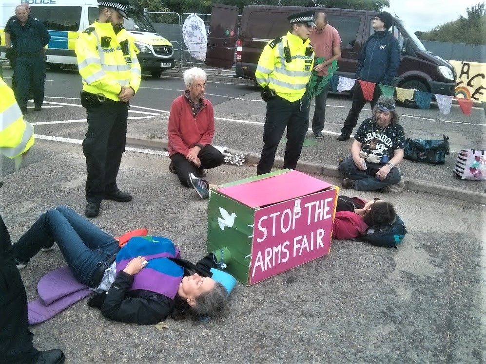 Image of campaigners "locked on" their arms joined and lying in a road. Two supporters are sat with them, three more standing behind. Police are present.